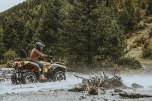 Person mit Helm und brauner Jacke fährt ATV Guardian 500 durch einen Fluss in bewaldeter Landschaft